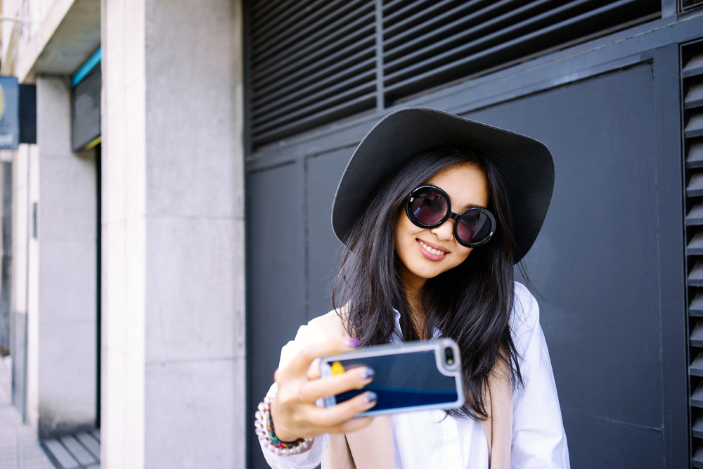 Young woman taking a selfie while wearing sunglasses and a black hat